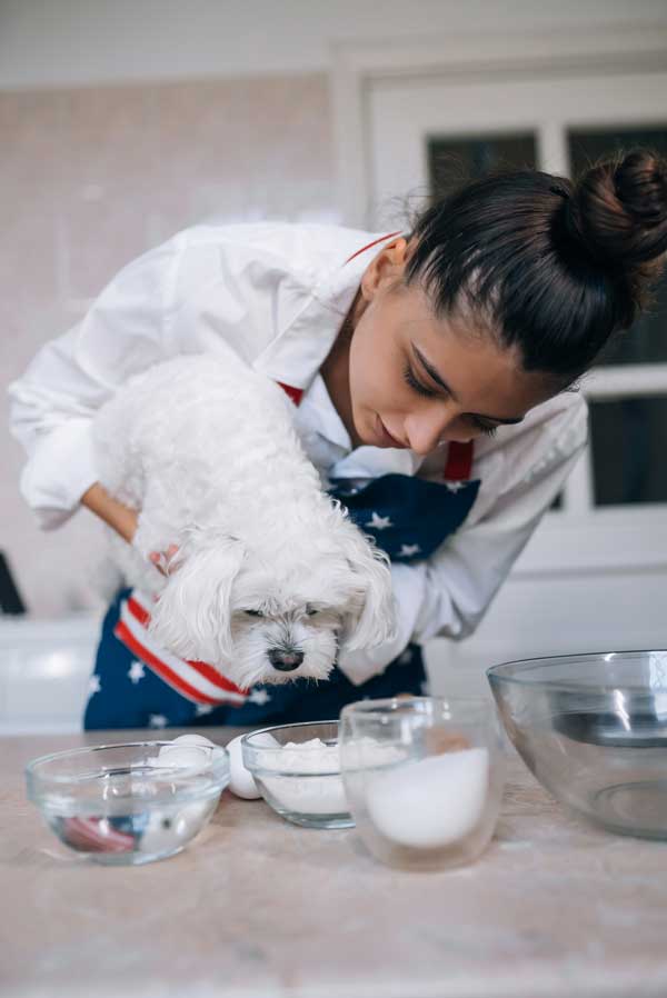 Woman holding a small white dog in a kitchen, examining ingredients, representing Rocker Dog’s canine nutritionist certified dog food recipes